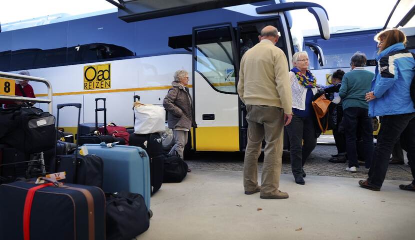 Oad-reizigers komen terug van hun busreis. Een groepje ouderen staat voor de bus, naast hen staan de koffers